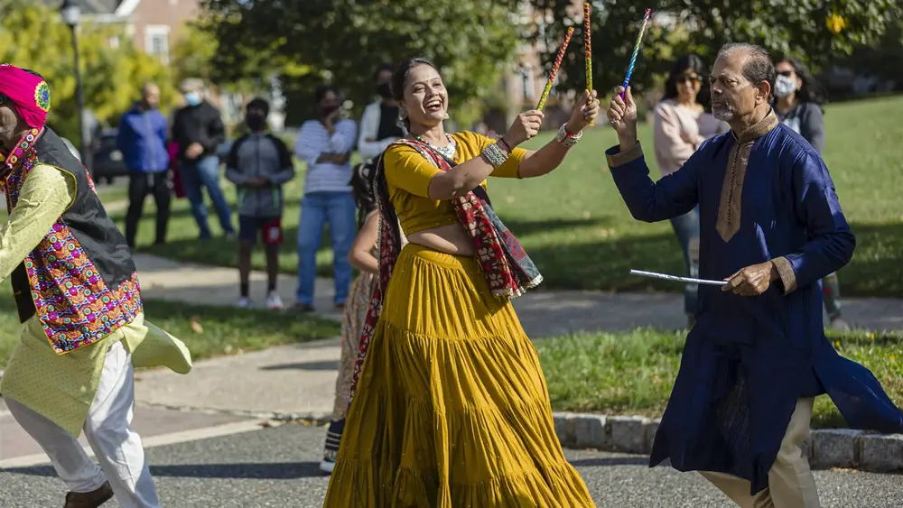 A woman in a yellow lehenga and a man in navy dance dandiya raas with sticks raised