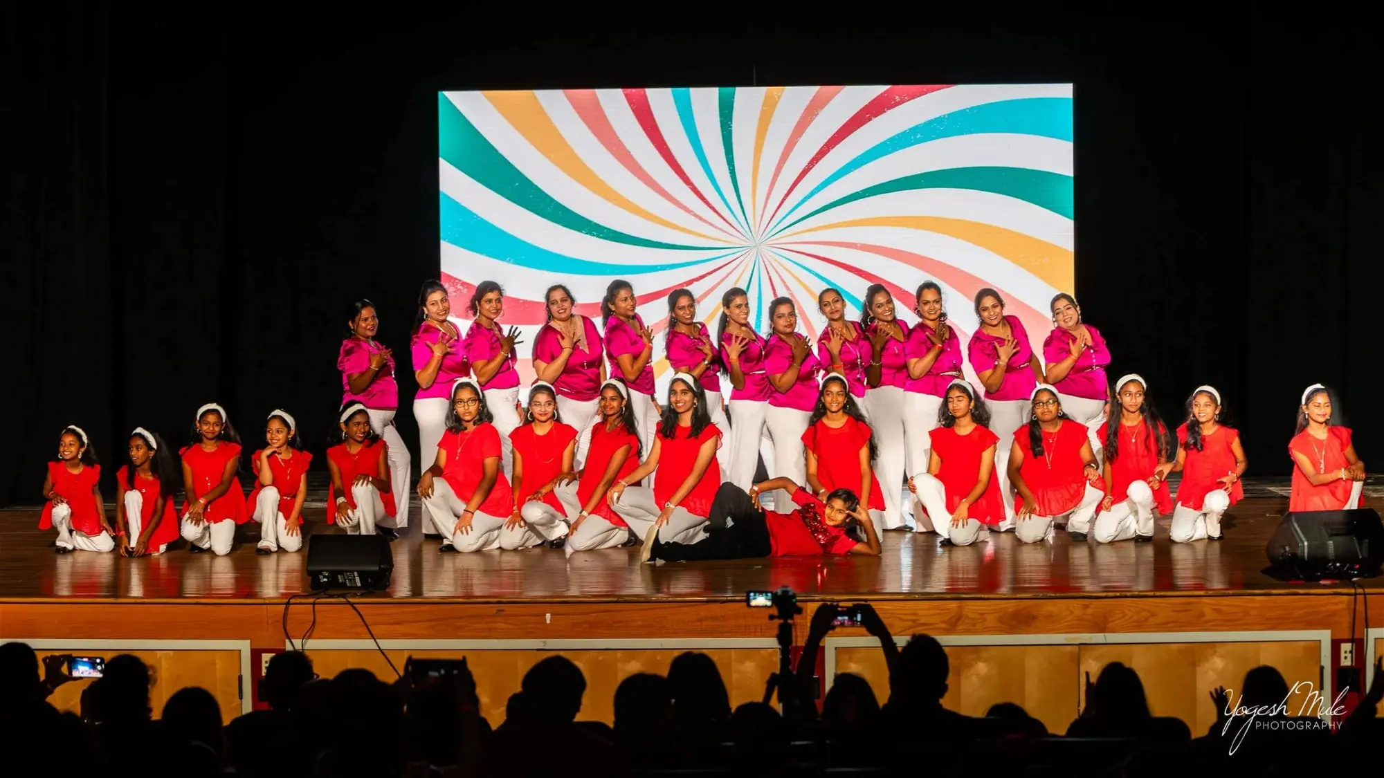 A chorus of children and adults in white and pink performs on the Diwali stage