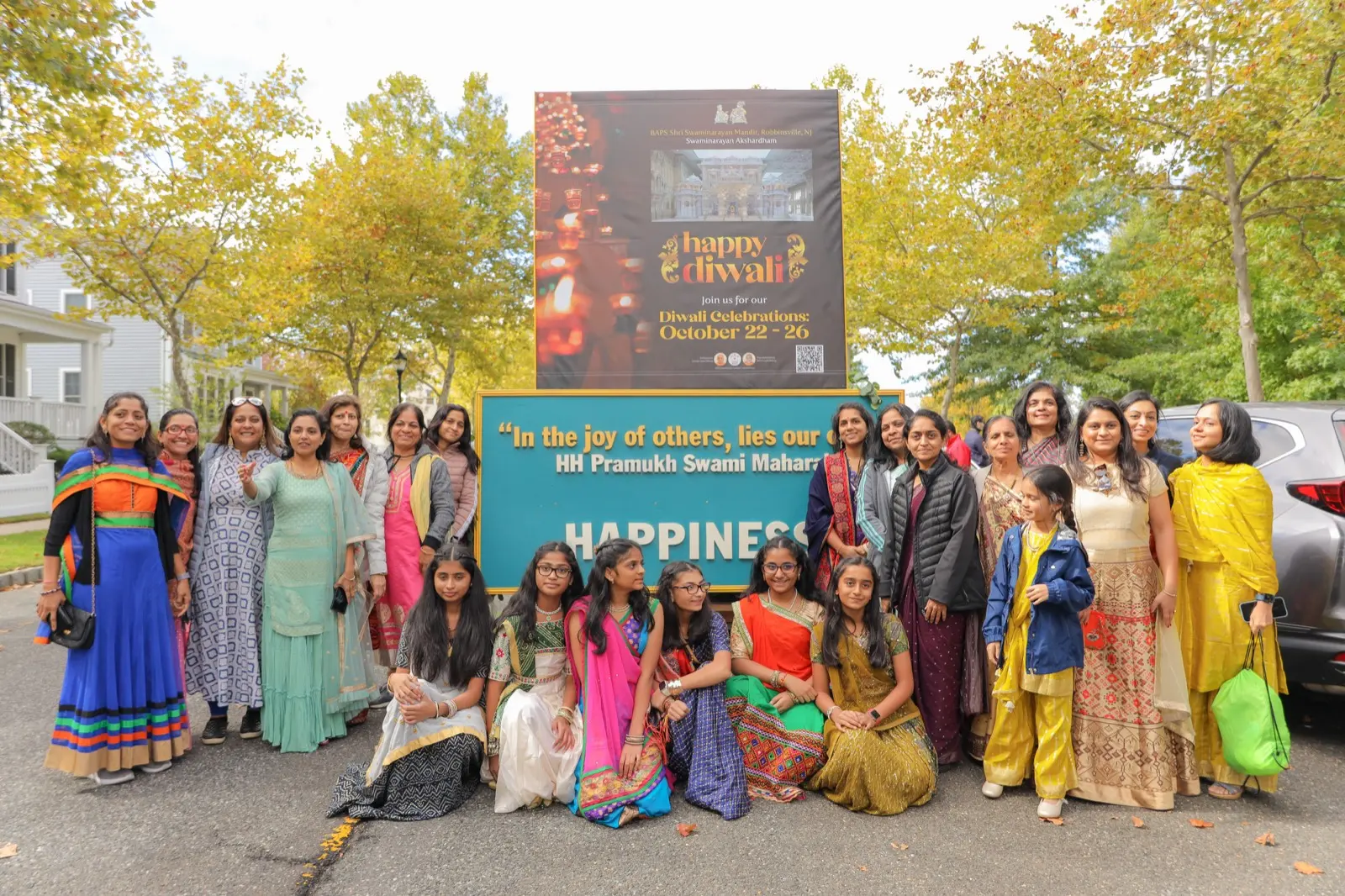 A Diwali parade contingent in festive clothing gathered behind its banner on a tree-lined street