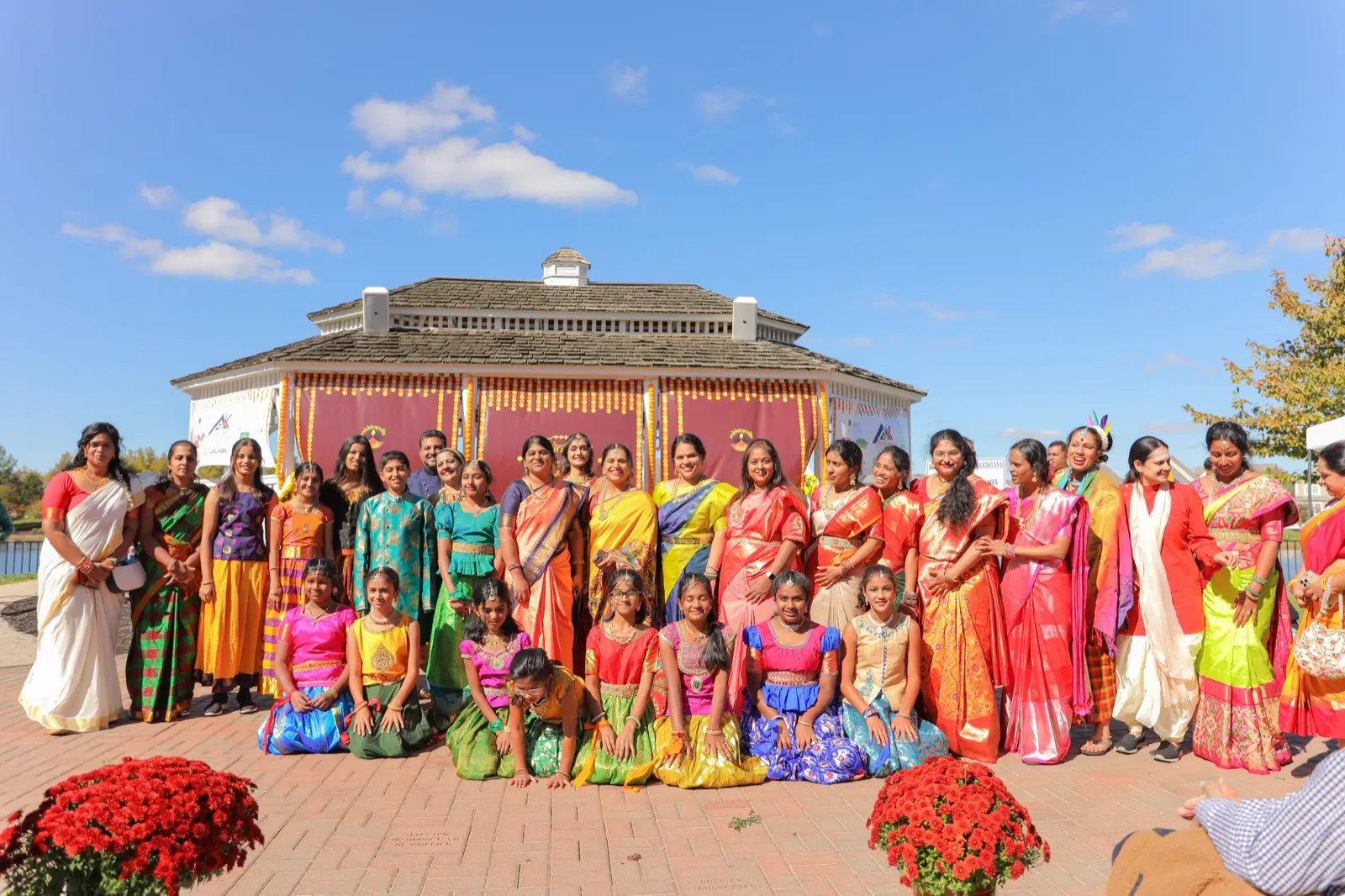 Performers in bright orange and pink costumes seated before the gazebo with marigold planters