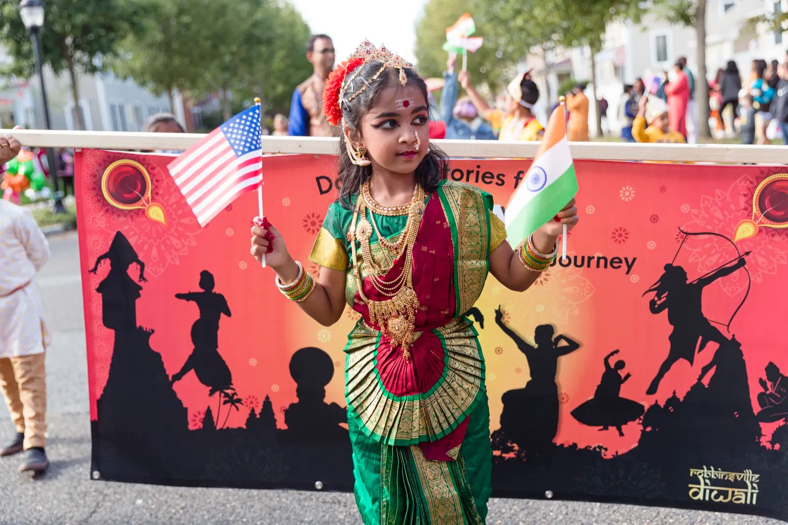 A young dancer in classical costume holds a small American flag at the Robbinsville Diwali Parade