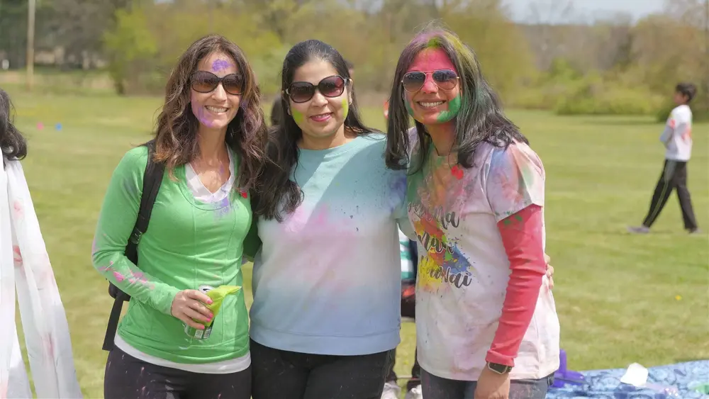 Three friends with pink and green colour on their faces smile at Robbinsville Holi
