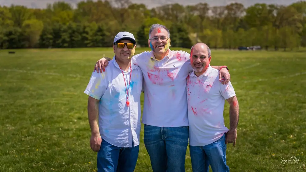 Three men in colour-splashed white shirts stand arm in arm at Holi