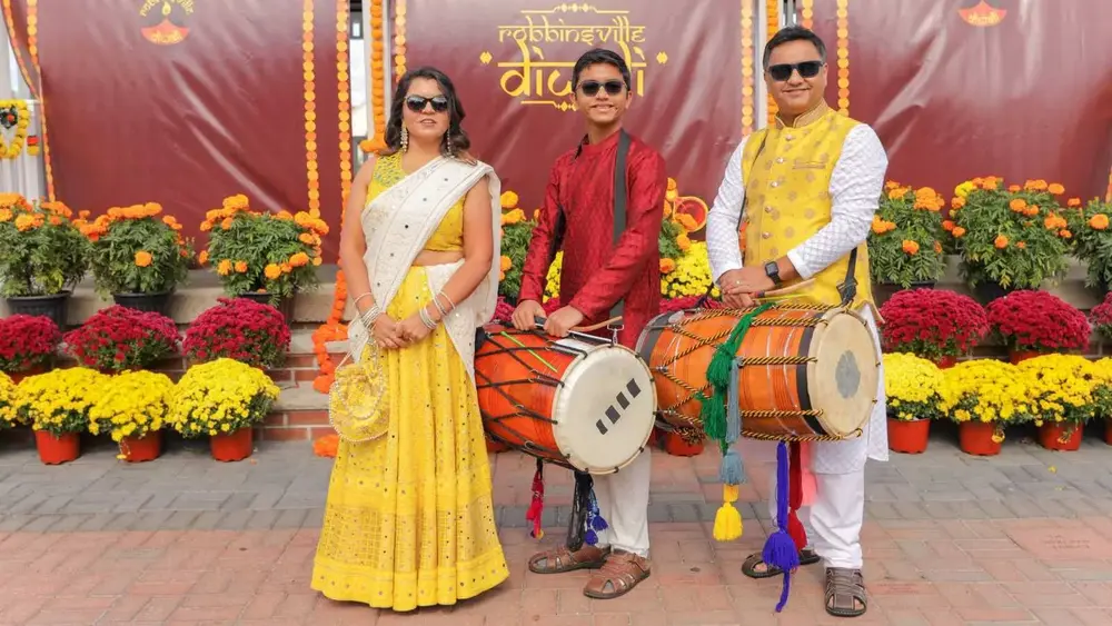 Two dhol drummers and a woman in a yellow lehenga in front of the Robbinsville Diwali backdrop and mums