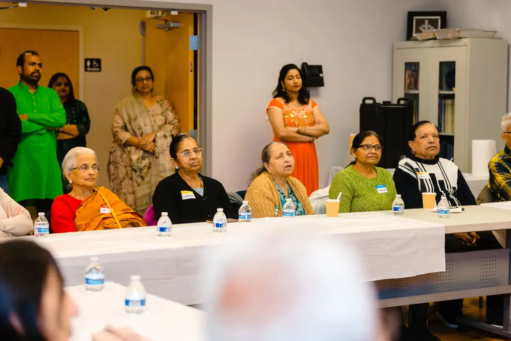 Seniors seated along a table smile toward the host at a Sanskriti gathering